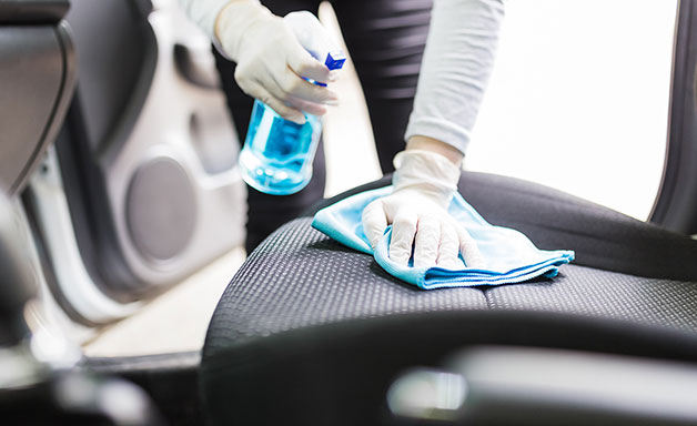 Close up of female wearing surgical gloves while cleaning car interior with disinfection liquid sprayer and microfiber cloth.